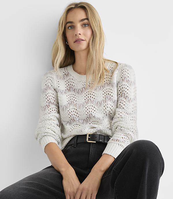 A blonde woman wearing a white crochet sweater and black jeans sits against a plain white background.