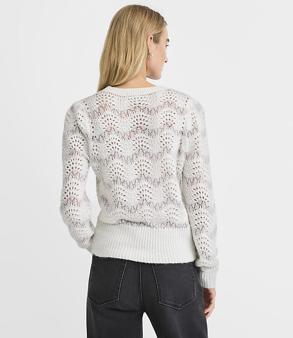 A woman wearing a white open-knit sweater and black jeans stands against a plain white background.
