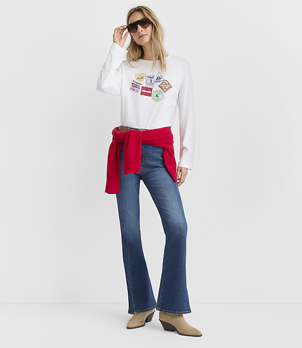 A woman wearing a white graphic long-sleeve shirt, blue flare jeans, and a red sweater tied around her waist stands against a plain white background.