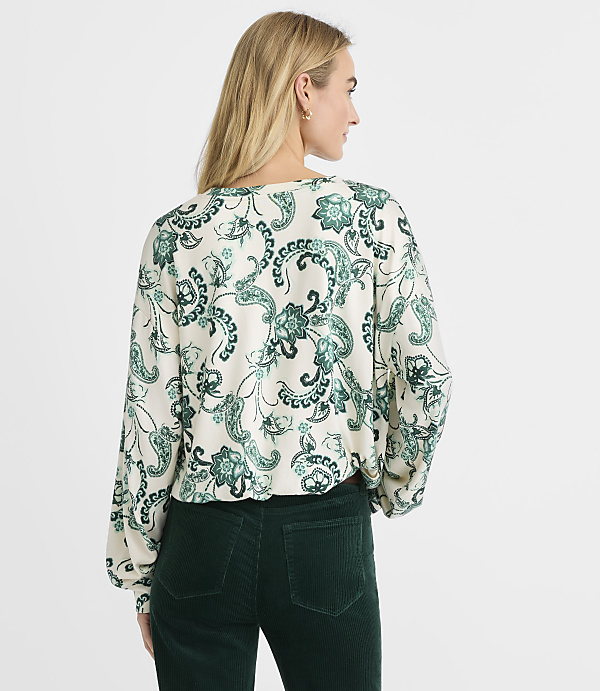 A woman wearing a green and white paisley print blouse and dark green corduroy pants stands against a plain white background.