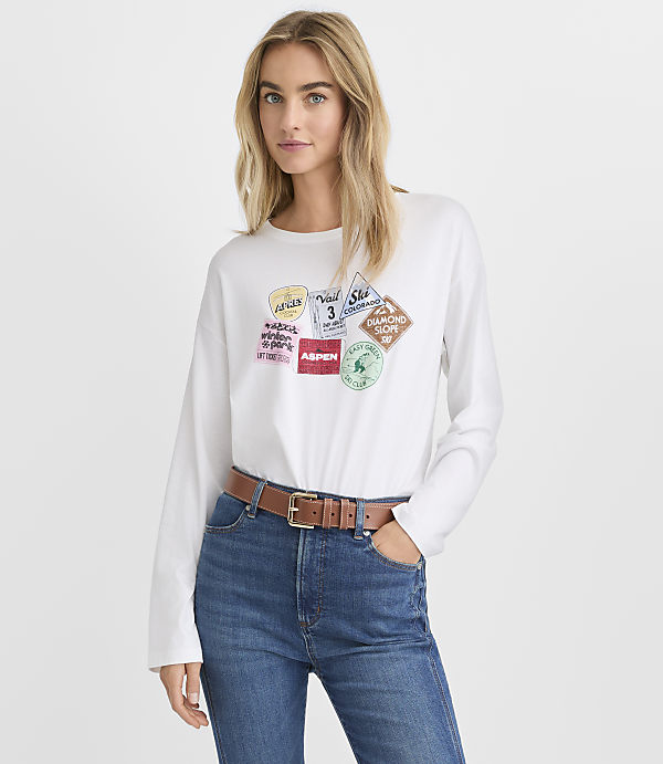 A blonde woman wearing a white long-sleeved graphic tee and blue jeans stands against a plain white background.