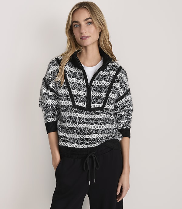 A woman wearing a patterned black and white quarter-zip sweater and black pants stands against a plain, light-colored background.