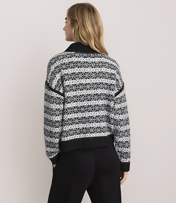 A woman with blonde hair wearing a patterned black and white sweater and black pants stands against a plain light-colored background.