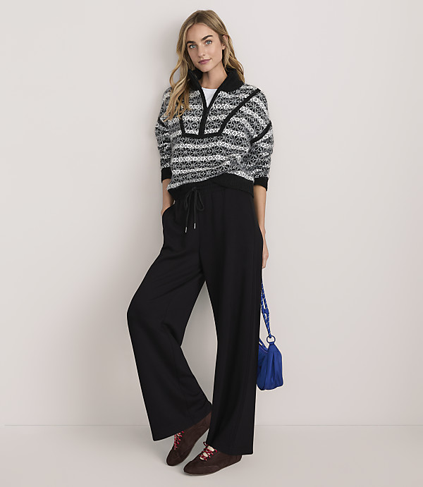 A woman wearing a patterned sweater and black wide-leg trousers stands against a plain, light-colored background.