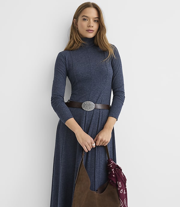 A woman wearing a long-sleeved blue turtleneck dress with a brown belt stands against a plain white background.