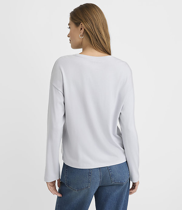 A woman wearing a light grey long-sleeved shirt and blue jeans stands with her back to the camera against a plain white background.