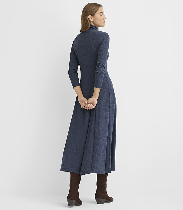 A woman wearing a long-sleeved navy blue maxi dress and brown boots stands with her back to the camera against a plain white background.