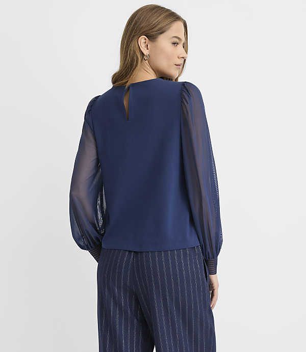 A woman wearing a navy blue blouse with sheer sleeves and pinstriped trousers stands against a plain white background.