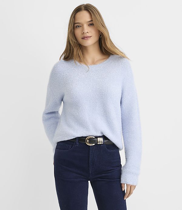 A woman wearing a light blue fuzzy sweater and dark blue corduroy pants stands against a plain white background.