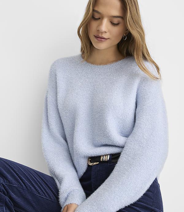 A woman wearing a light blue fuzzy sweater and dark blue corduroy pants poses against a plain white background.