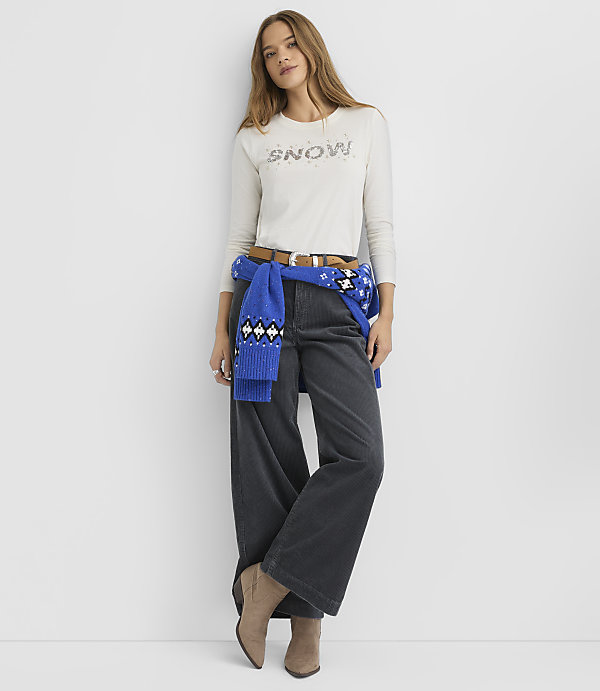 A woman wearing a white "SNOW" shirt, wide-leg dark trousers, and a blue sweater tied around her waist stands against a plain white background.