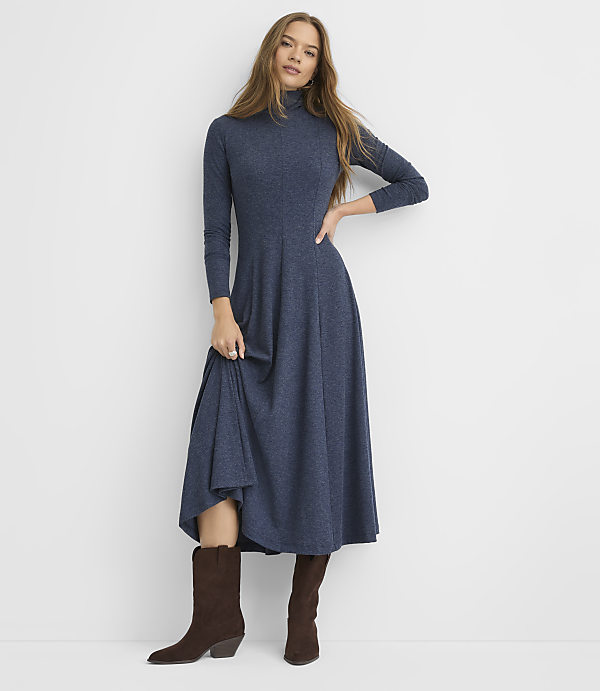 A woman wearing a long-sleeved blue maxi dress and brown boots stands against a plain white background.