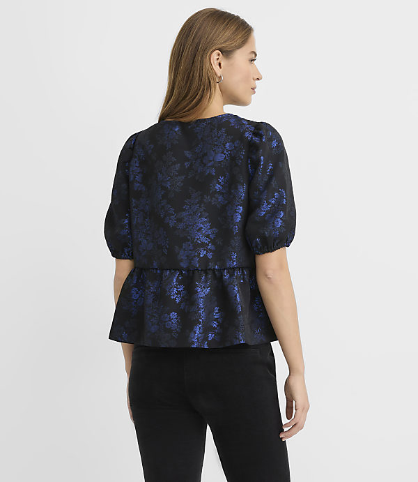 A woman wearing a blue floral peplum top and black pants stands against a plain white background.