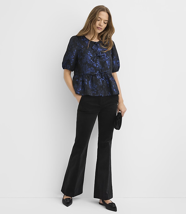 A woman wearing a blue floral blouse and black flared trousers stands against a plain white background.