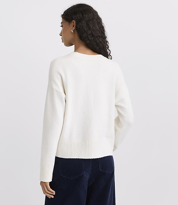 A woman wearing a white sweater and dark blue trousers stands against a plain light gray background.