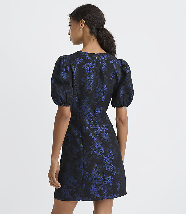A woman stands with her back to the camera wearing a short, puff-sleeved blue and black floral dress against a plain white background.
