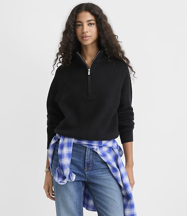A woman wearing a black quarter-zip sweater and blue jeans with a plaid shirt tied around her waist stands against a plain white background.