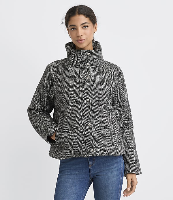 A woman wearing a grey and black herringbone patterned jacket and blue jeans stands against a plain white background.