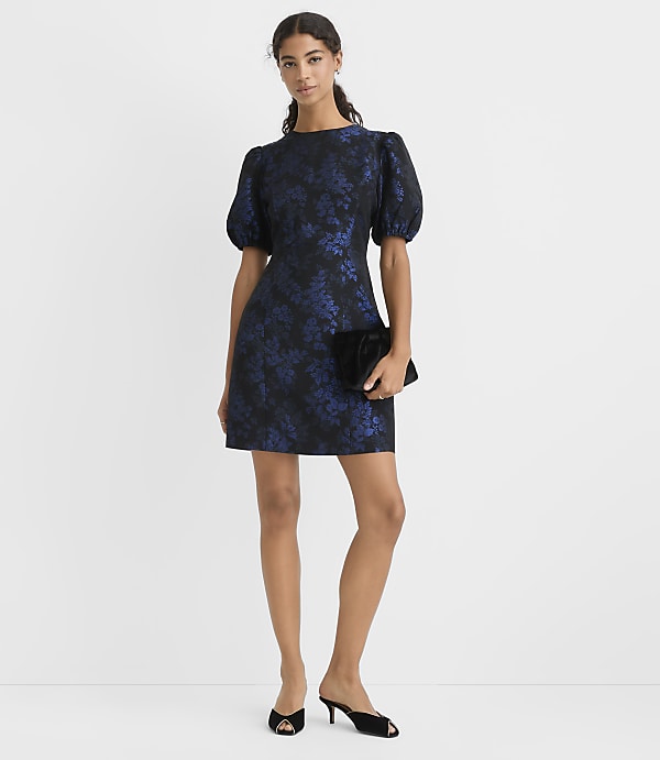 A woman wearing a navy blue floral puff-sleeve dress and black heels stands against a plain white background.