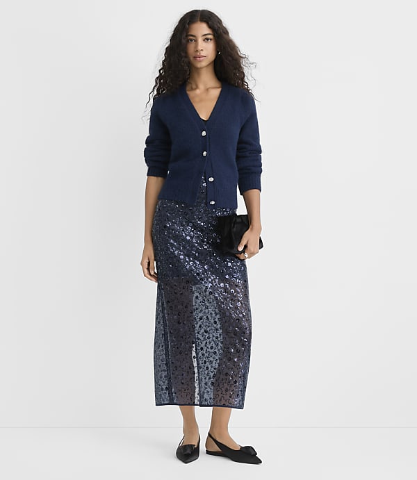 A woman wearing a navy blue cardigan and a shimmering sequined skirt stands against a plain white background.