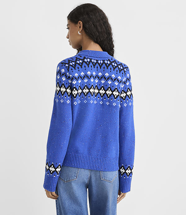 A woman wearing a blue patterned sweater and jeans stands against a plain white background.