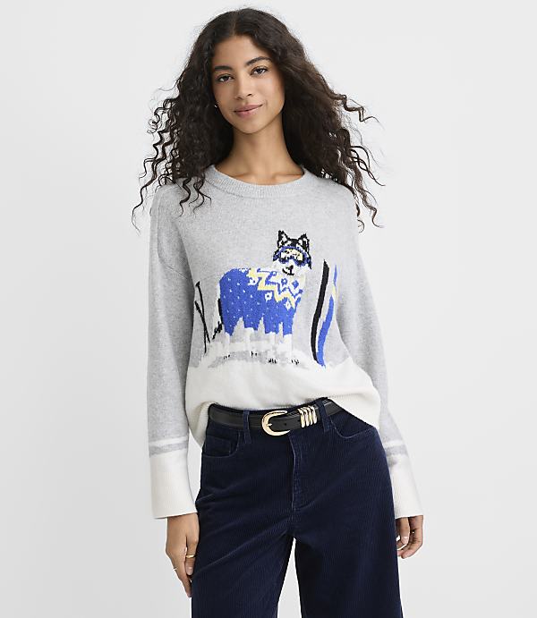 A woman with curly hair wears a grey and white sweater with a blue llama graphic and dark blue corduroy pants against a plain white background.