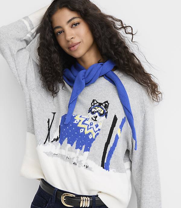 A woman with long curly hair wears a grey and white patterned sweater with a blue scarf against a plain white background.
