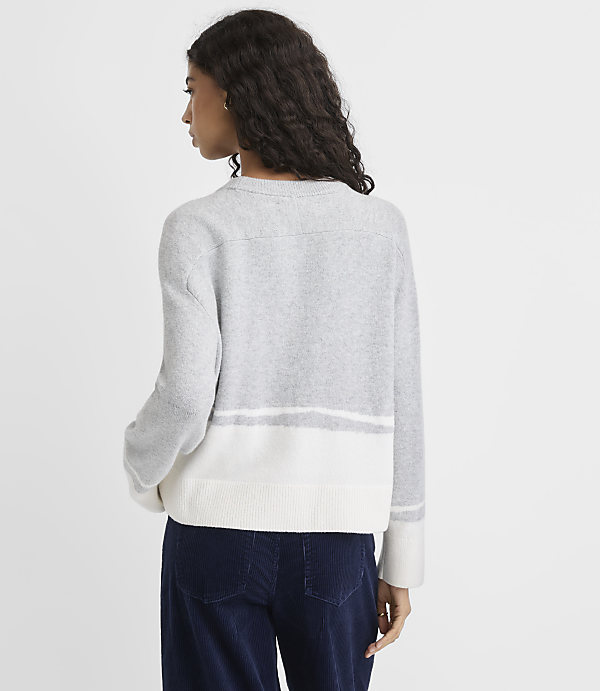 A woman wearing a grey and white striped sweater and navy blue pants stands against a plain light grey background.
