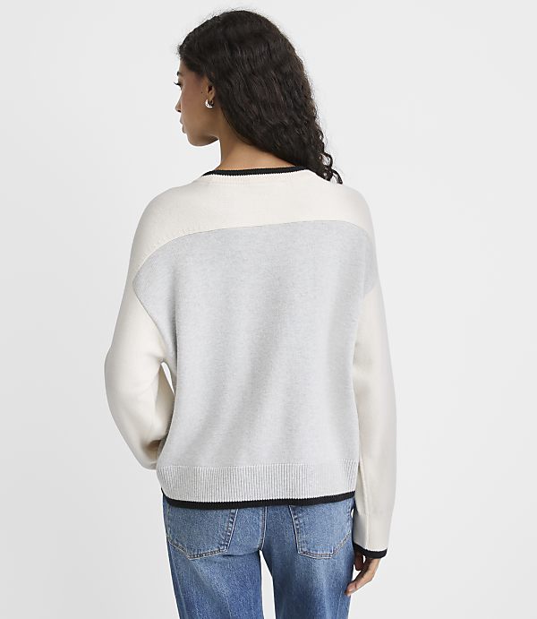 A woman wearing a color-blocked white and grey sweater and blue jeans stands against a plain light grey background.