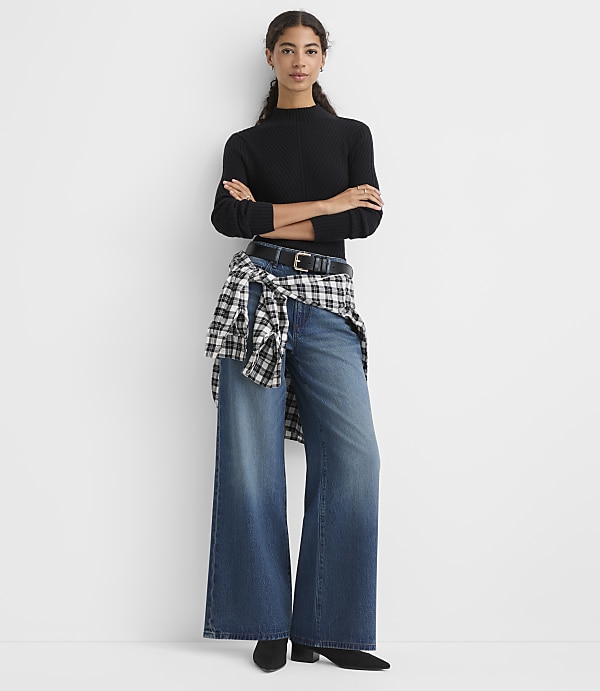 A woman wearing a black ribbed top, wide-leg jeans, and a checkered shirt tied around her waist stands against a plain white background.