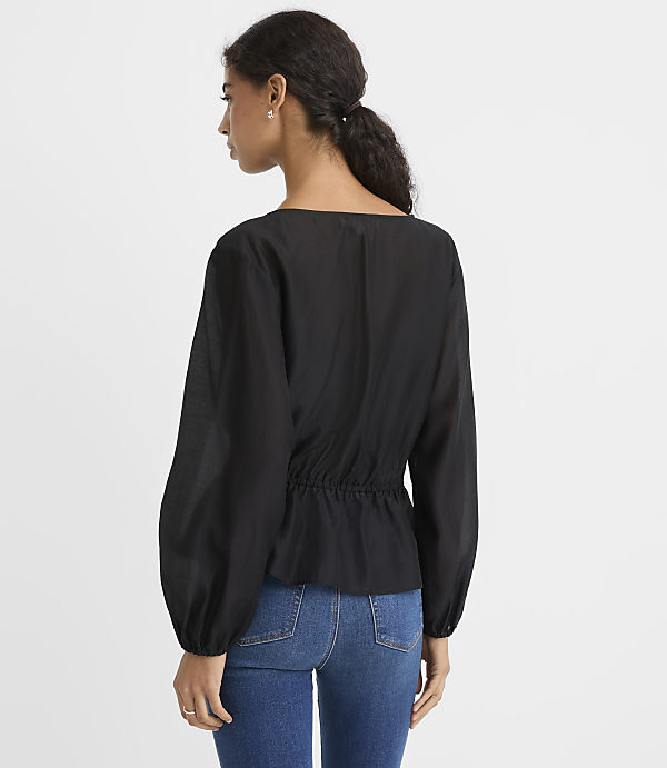 A woman wearing a black long-sleeved blouse and blue jeans stands against a plain white background.