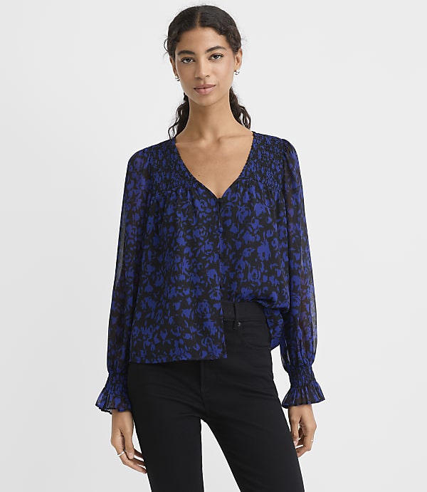 A woman wearing a blue and black floral print blouse and black pants stands against a plain white background.