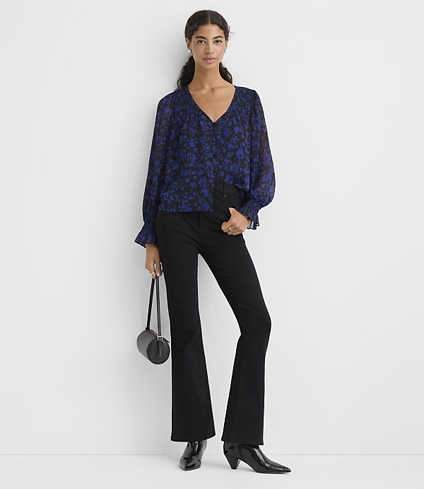 A woman wearing a blue floral blouse and black flared trousers stands against a plain white background.