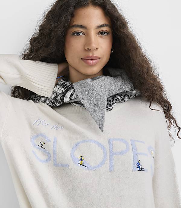 A woman with long curly hair wearing a white graphic sweater and a grey patterned scarf poses against a plain white background.