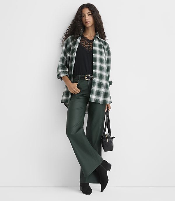A woman wearing a green plaid shirt, black top, and dark green trousers stands against a plain white background.