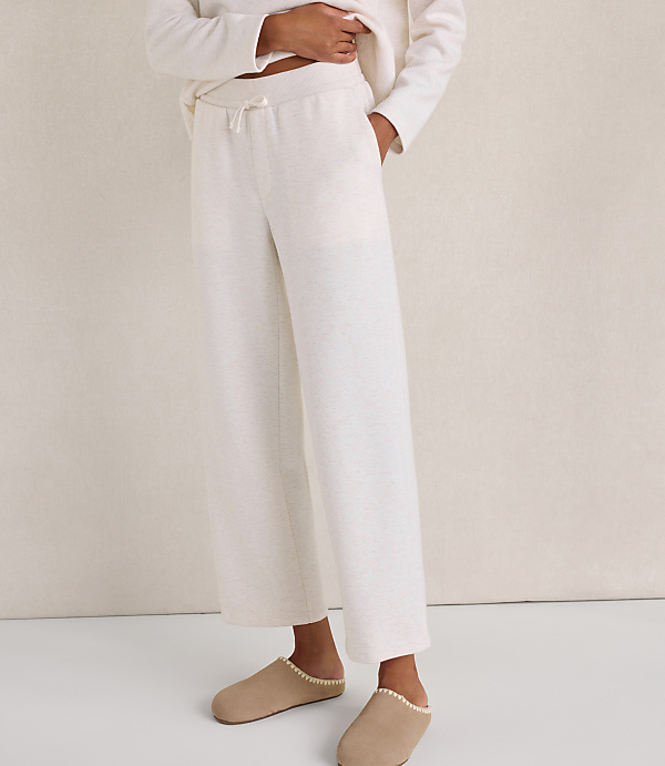 A person wearing cream-colored lounge pants and beige slippers stands against a plain, neutral-toned wall.