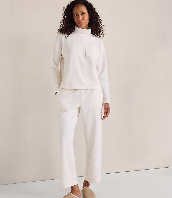 A woman wearing a cream-colored lounge set and slippers stands against a plain, neutral-toned background.