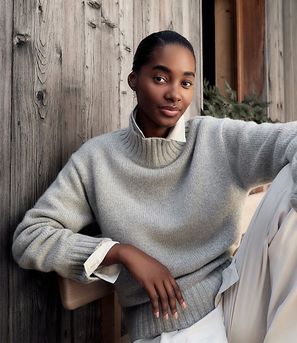 A woman wearing a grey sweater and white collared shirt sits against a rustic wooden wall.
