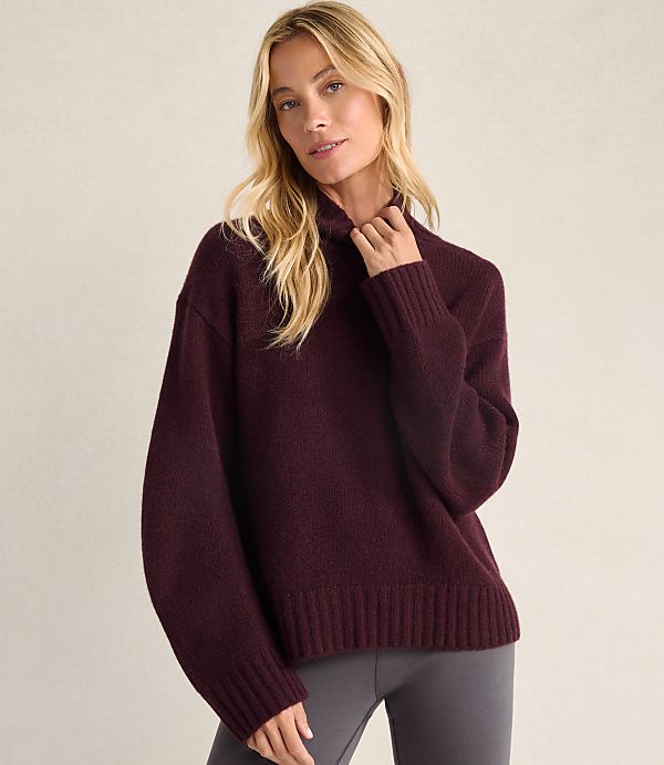 A blonde woman wearing a maroon oversized sweater and grey leggings stands against a plain, light-colored background.