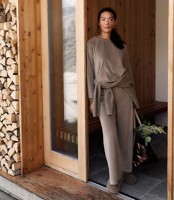 A woman in a taupe lounge set stands in a wooden doorway, with a stack of firewood to her left and a rustic interior behind her.