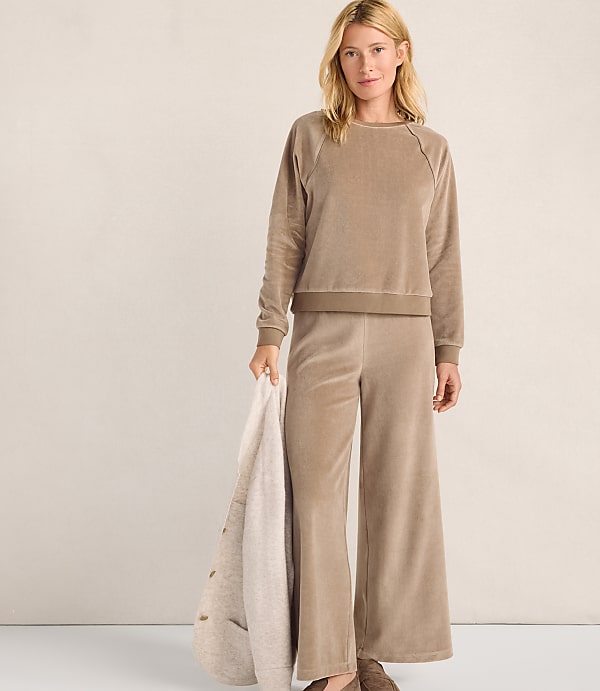 A woman wearing a tan velvet lounge set stands against a plain, light-colored wall while holding a cream-colored blanket.