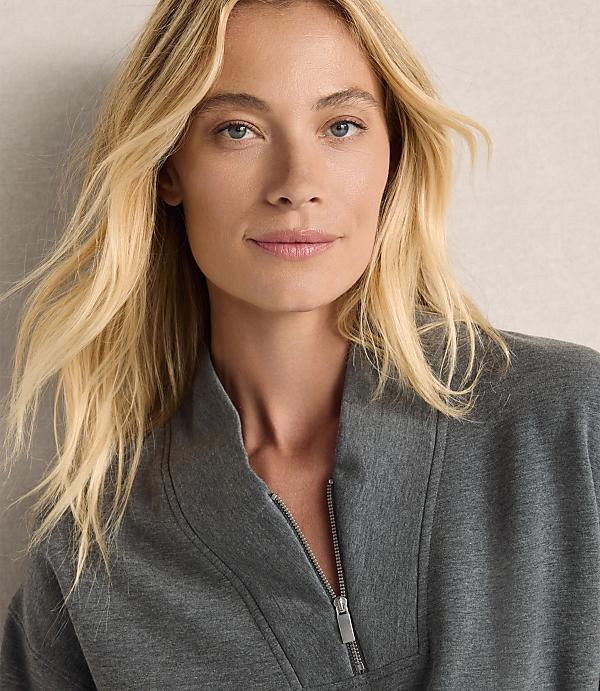 A blonde woman wearing a grey zip-up jacket stands against a plain, neutral-toned background.