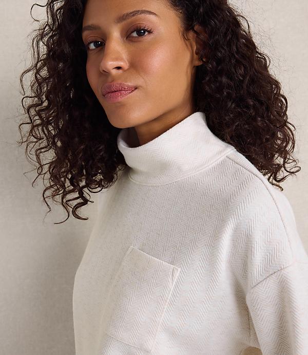A woman with curly dark hair wearing a white textured turtleneck sweater stands against a plain, light-colored wall.