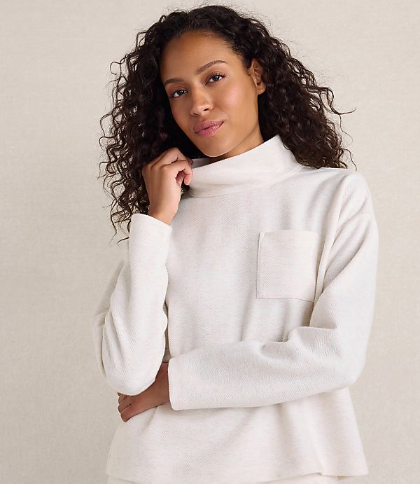 A woman with curly dark hair wearing a white textured turtleneck sweater poses against a plain beige background.