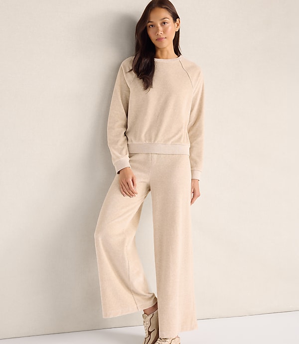 A woman wearing a beige long-sleeved top and matching wide-leg trousers stands against a plain, light-colored wall.
