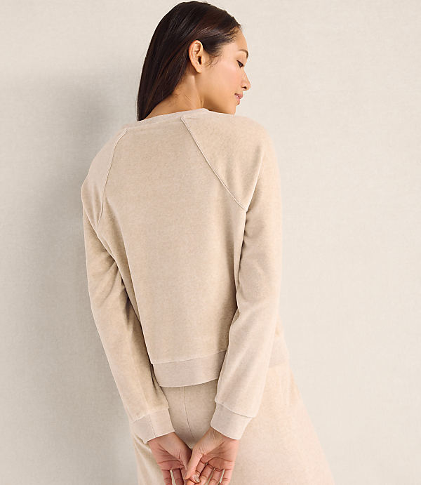 A woman wearing a beige sweatshirt and matching pants stands with her back to the camera against a plain, light-colored wall.