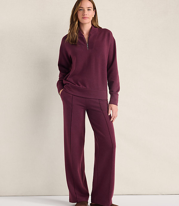 A woman wearing a maroon quarter-zip pullover and matching wide-leg pants stands against a plain, light-colored wall.