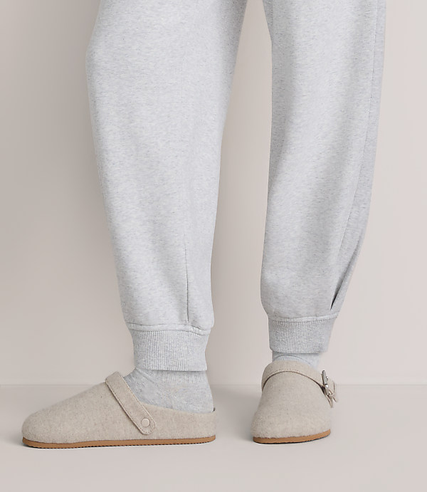 A person wearing light grey sweatpants and beige slippers stands against a plain, neutral-colored background.