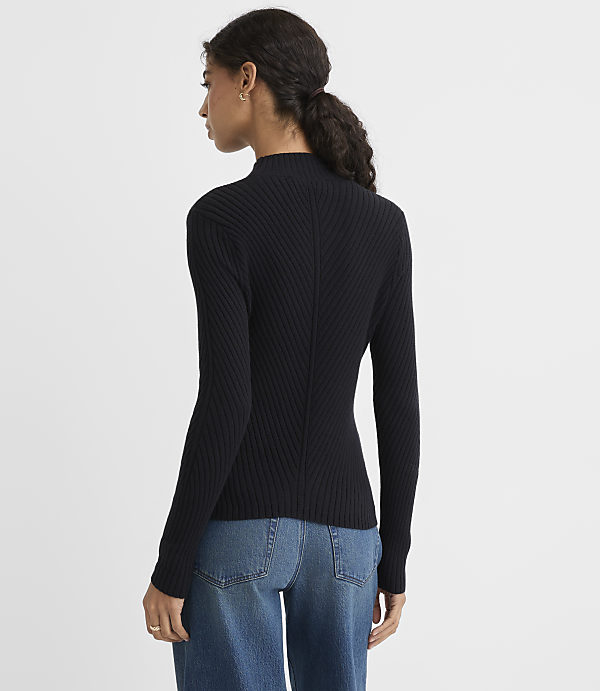 A woman wearing a black ribbed turtleneck and blue jeans stands against a plain white background.