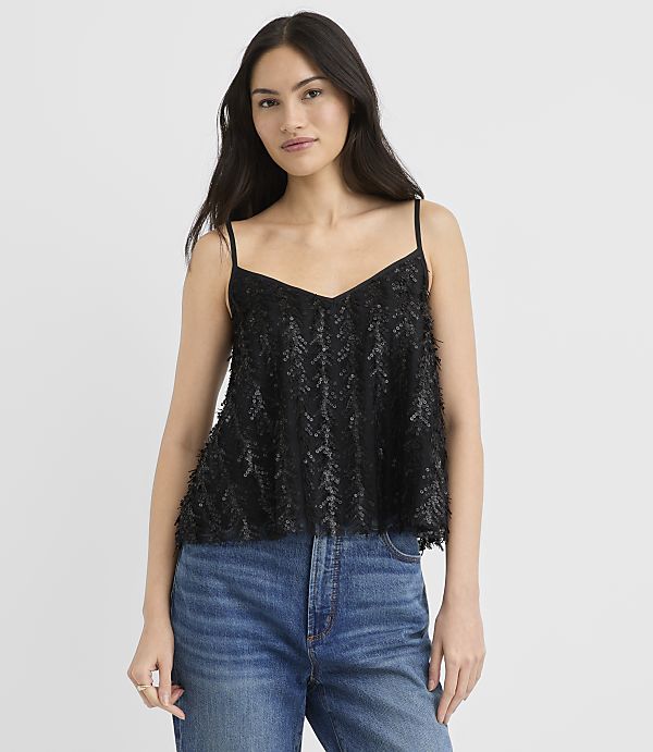 A woman wearing a black sequined camisole and blue jeans stands against a plain white background.